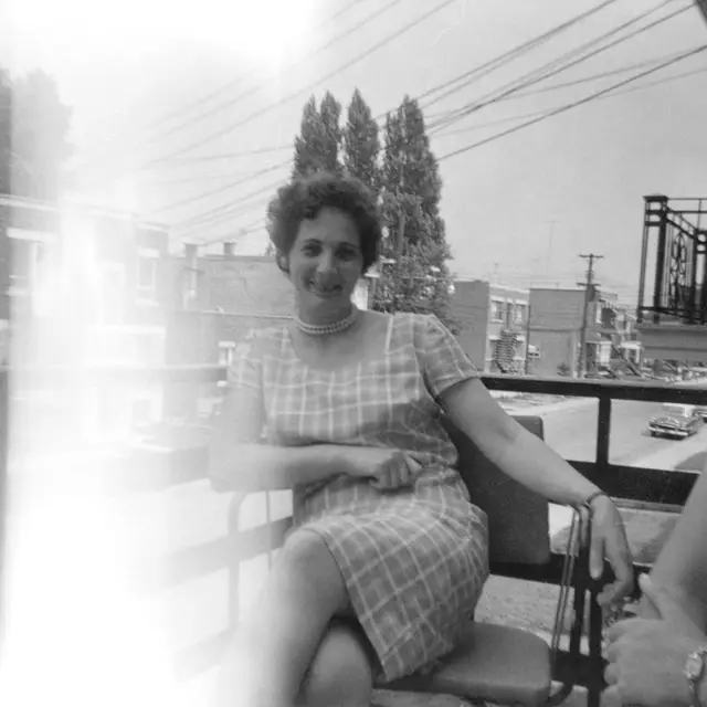 Jeanne, jeune, assise et souriante sur un balcon, en robe à carreaux.